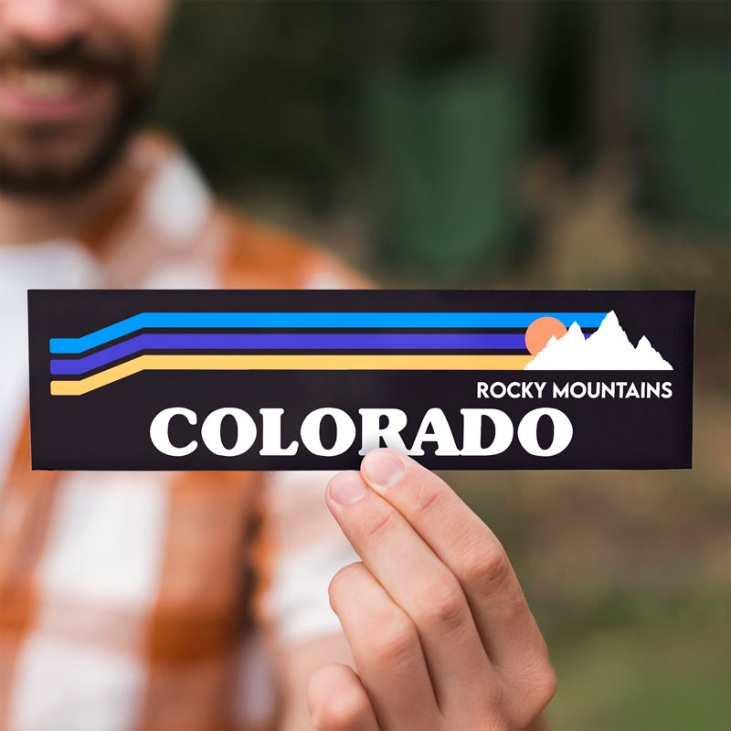 Person holding a Colorado bumper sticker with a retro mountain design and 'Rocky Mountains' text, set against a blurred outdoor background.