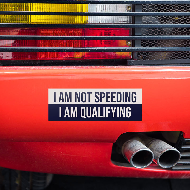 Red sports car with a humorous bumper sticker reading 'I Am Not Speeding, I Am Qualifying,' placed near the exhaust pipes.