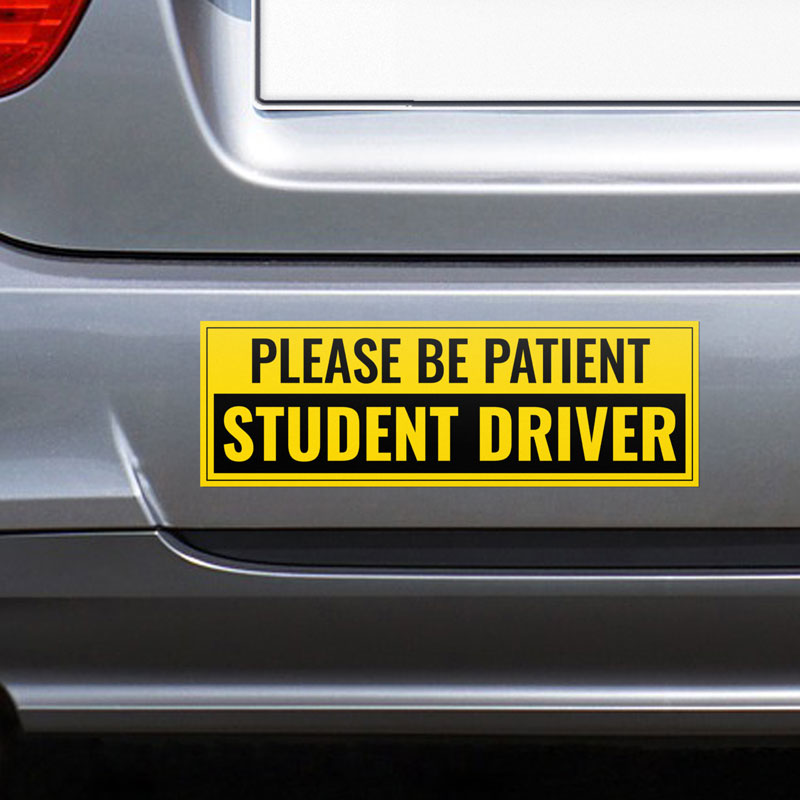 Silver car with a yellow 'Please Be Patient, Student Driver' bumper sticker in bold black text for road safety awareness.