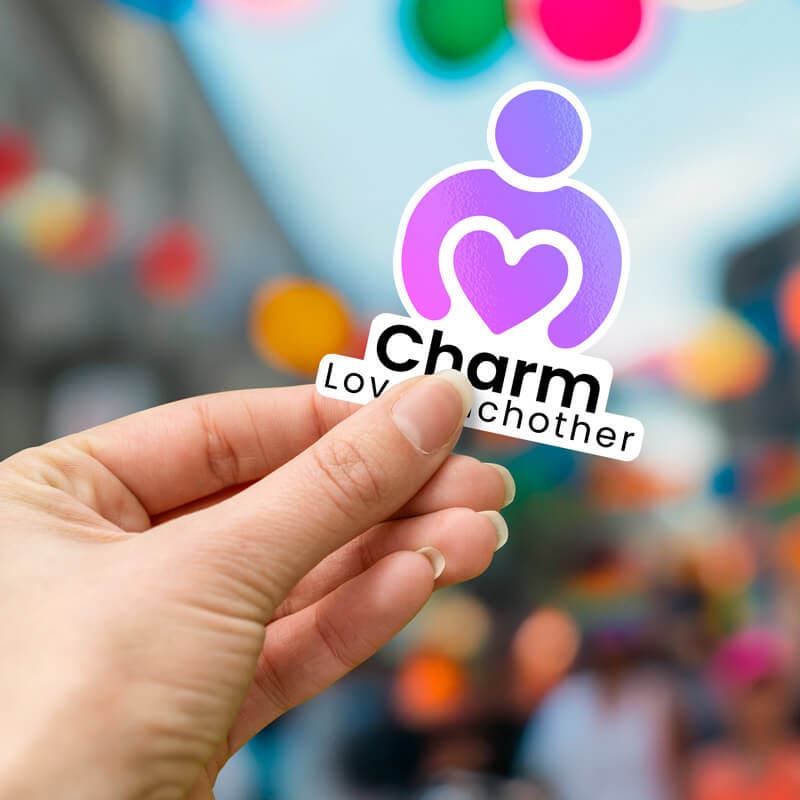 Hand holding a 'Charm Love Each Other' sticker with a purple heart design, against a blurred background with colorful lights.