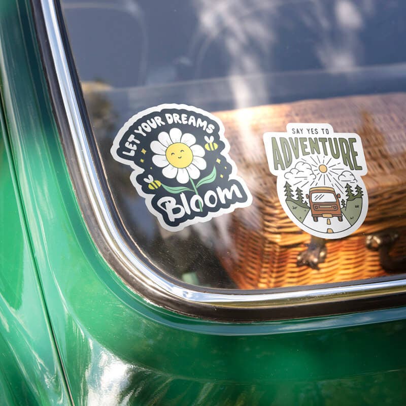 Car window with 'Let Your Dreams Bloom' and 'Say Yes to Adventure' stickers, featuring a daisy and a retro van design.