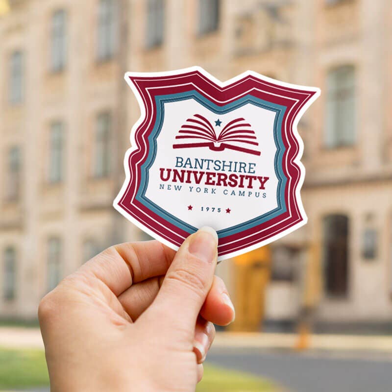 Hand holding a university crest sticker with 'Bantshire University' logo in red and blue, set against a blurred campus background.