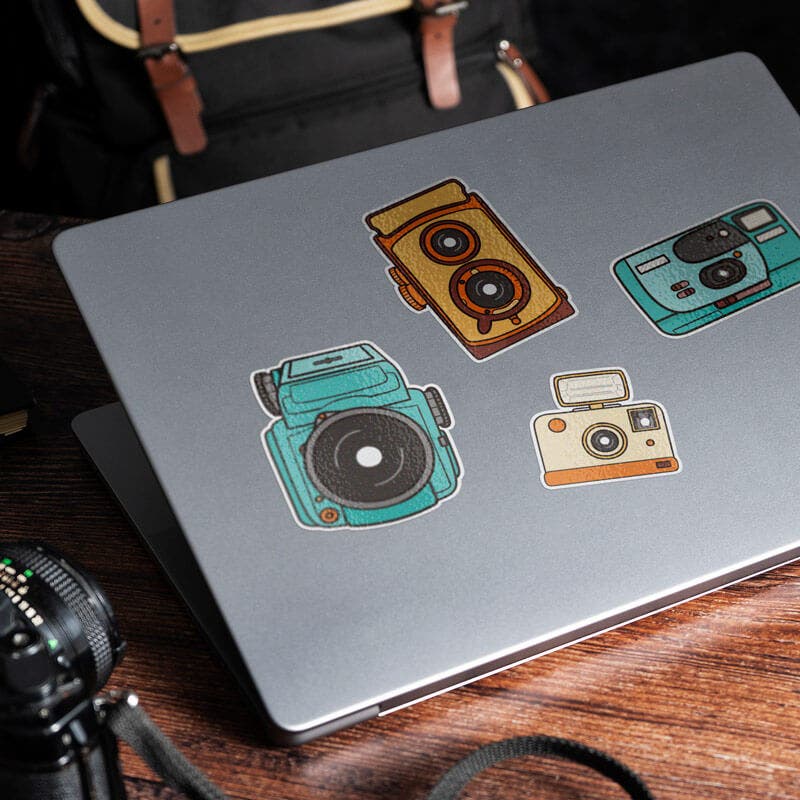 Gray laptop decorated with vintage camera stickers in various colors, sitting on a wooden desk with a retro photography theme.