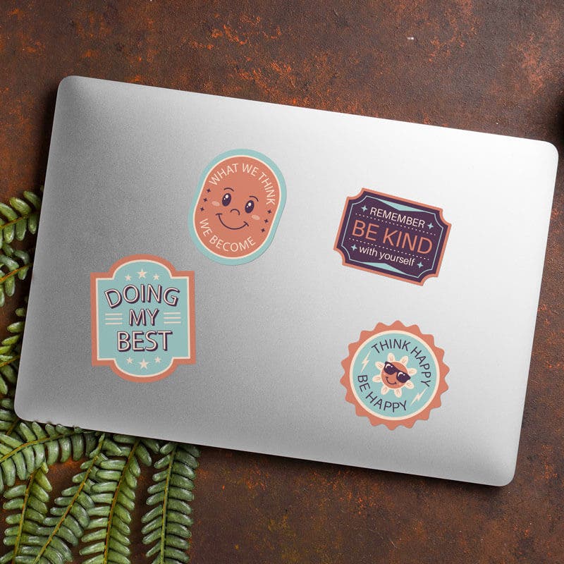 Gray laptop adorned with motivational stickers, including 'Doing My Best' and 'Be Kind,' resting on a textured brown surface with ferns.