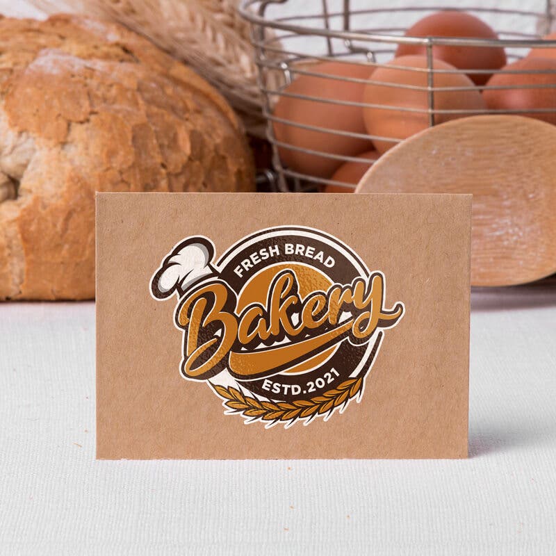 A brown paper card featuring a bakery logo sticker with a wheat and bread design, placed on a table with fresh bread, eggs, and a basket in the background.