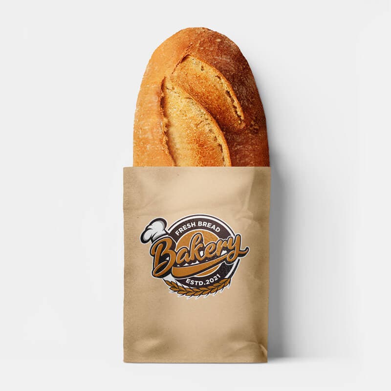 A loaf of bread in a paper bag featuring a bakery logo sticker with a wheat and bread motif, placed against a plain white background.