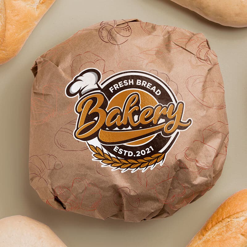 A round loaf of bread wrapped in brown paper with a bakery logo sticker, surrounded by other freshly baked goods on a neutral background.