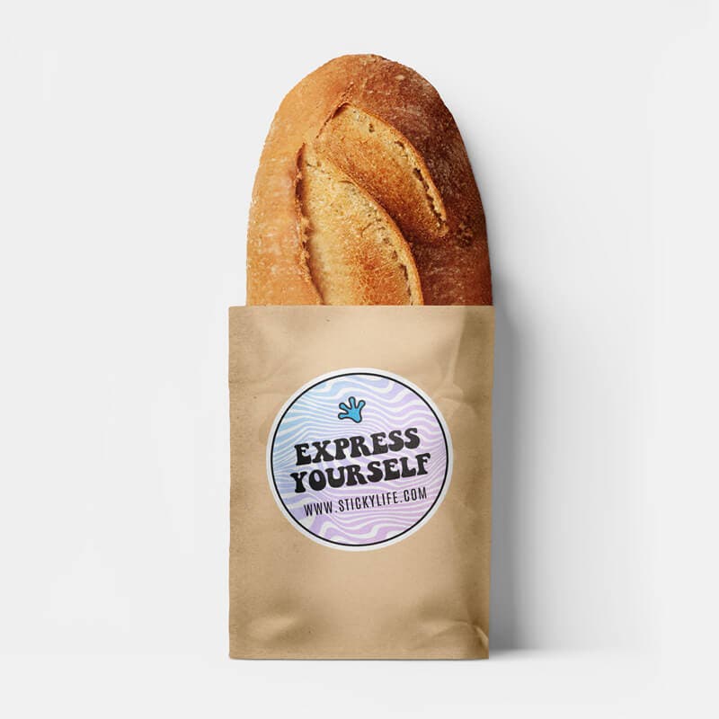 A loaf of bread inside a brown paper bag featuring a bakery logo sticker with a pastel wavy design, set against a plain white background.